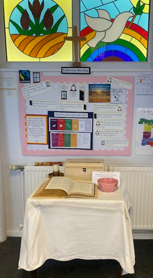 Picture shows the Collective Worship display in the entrance hall. In the background are stained glass windows with pictures reflecting school values of Hope and Peace. On the table is a Bible, prayer box and  prayer beads.