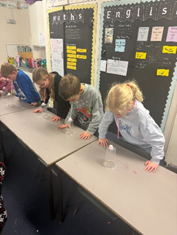 A group of children hovering over a plastic with a marshmallow on top. The children are trying to get the marshmallow off with a straw.  