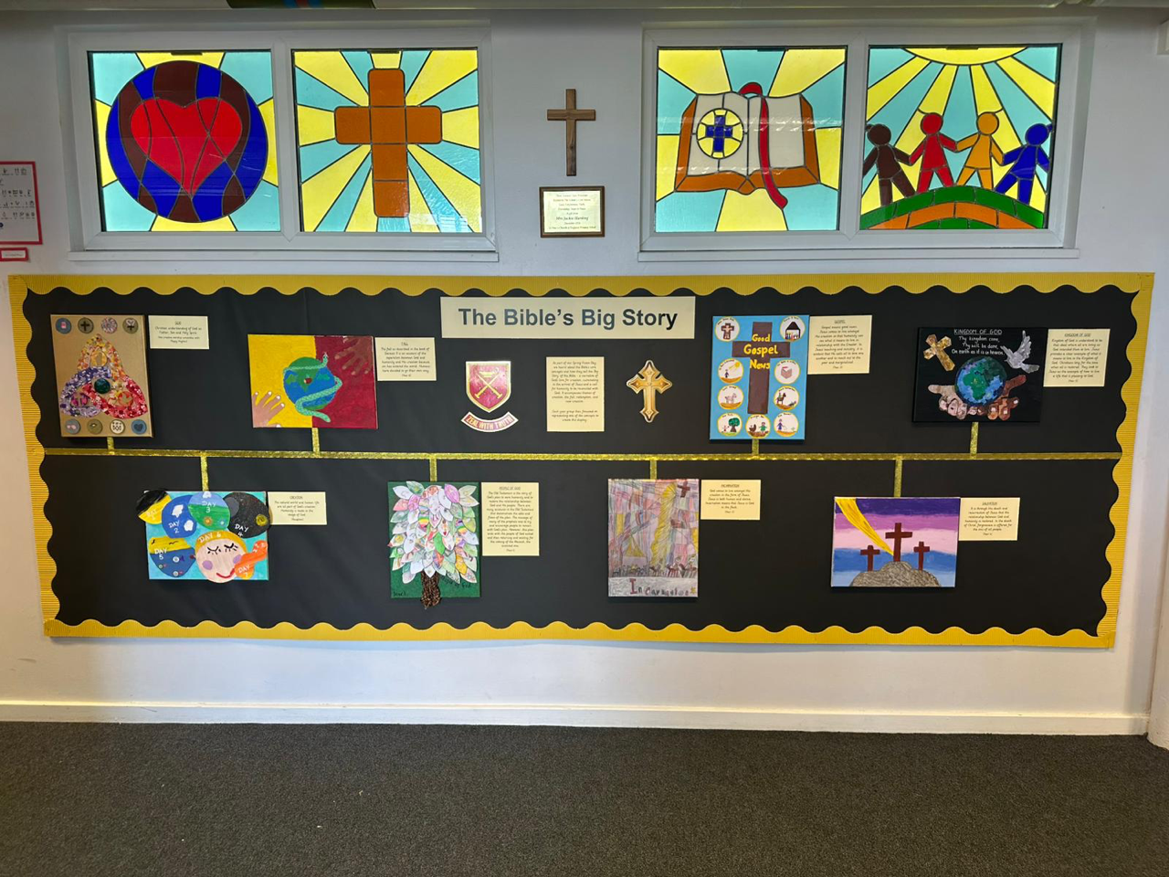 Image shows a display board backed in black featuring a colourful A3 artwork produced by each Year Group arranged on a timeline to illustrate the Core Concepts of the Bible. The title of the board is ‘The Bible’s Big Story’. Above the board are stained glass windows illustrating the school’s values of love, faith, forgiveness and friendship.