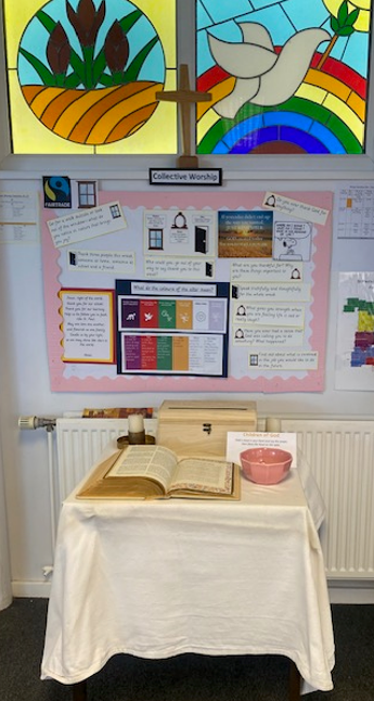 Picture shows the Collective Worship display in the entrance hall. In the background are stained glass windows with pictures reflecting school values of Hope and Peace. On the table is a Bible, prayer box and  prayer beads.