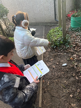 A child pointing at a carrot on the floor and another child logging the carrot on their data sheet. 